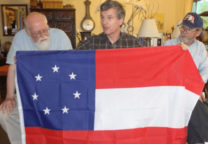 At the July 2015 PFA meeting, Ted Kaye provides a summary of the flags of the Confederacy, displaying the first national flag, the “Stars and Bars” as John Schilke and Fred Paltridge look on.